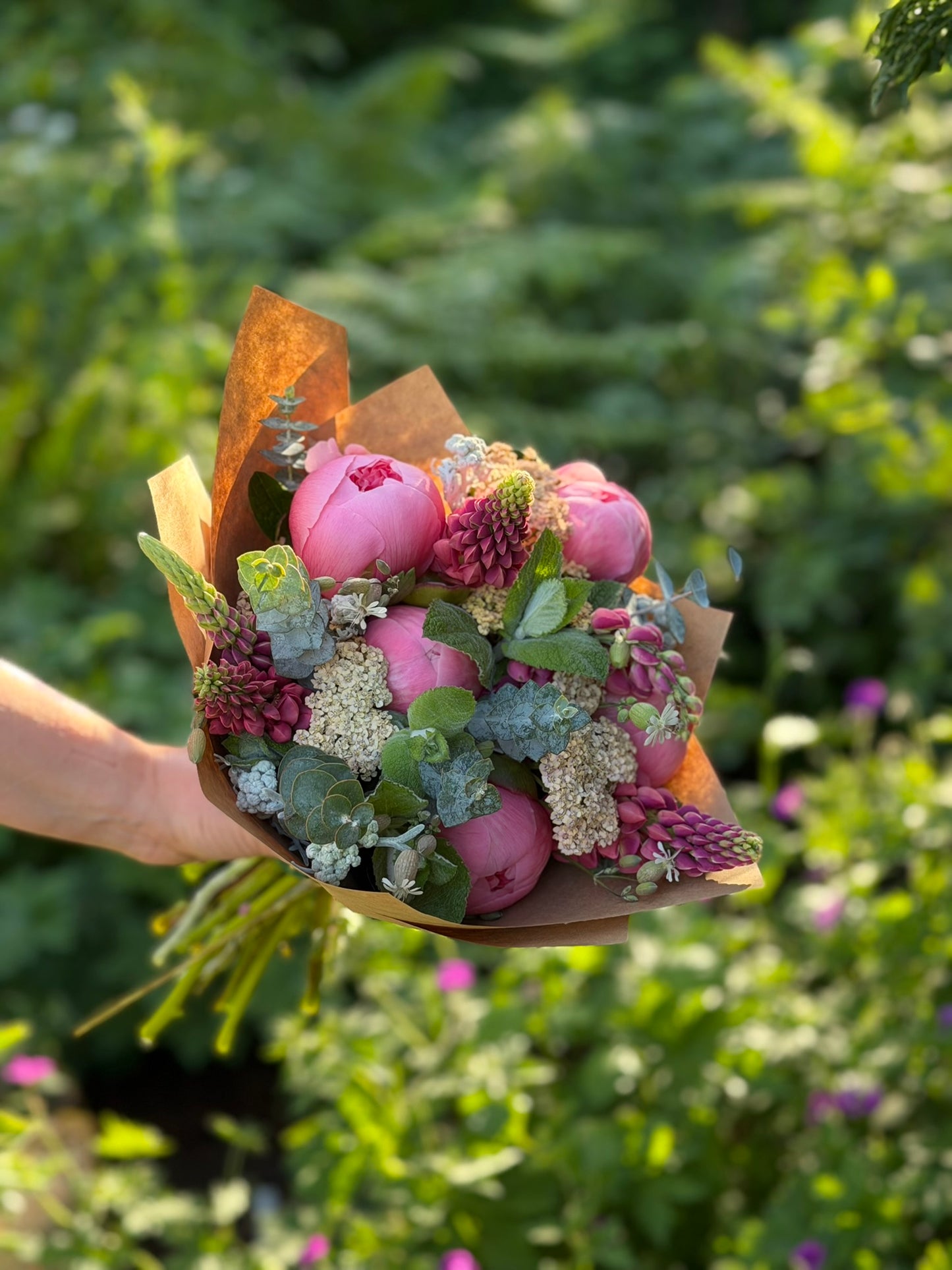 A hand holds a graceful-stem Bouquets & Arrangements for Delivery, featuring pink peonies and greenery wrapped in brown paper, set against a blurred garden background.