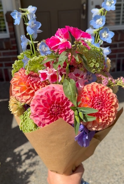 A hand holds the Gracefully Hand-Tied Bouquet from The Graceful Stem, featuring an assortment of fresh flowers such as pink dahlias, blue delphiniums, pink carnations, and green foliage, all beautifully wrapped in brown paper.