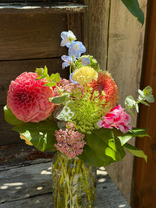 A Graceful Mason arrangement by The Graceful Stem, filled with hand-designed blooms and greenery, sits on a wooden surface in sunlight against a rustic wooden background.
