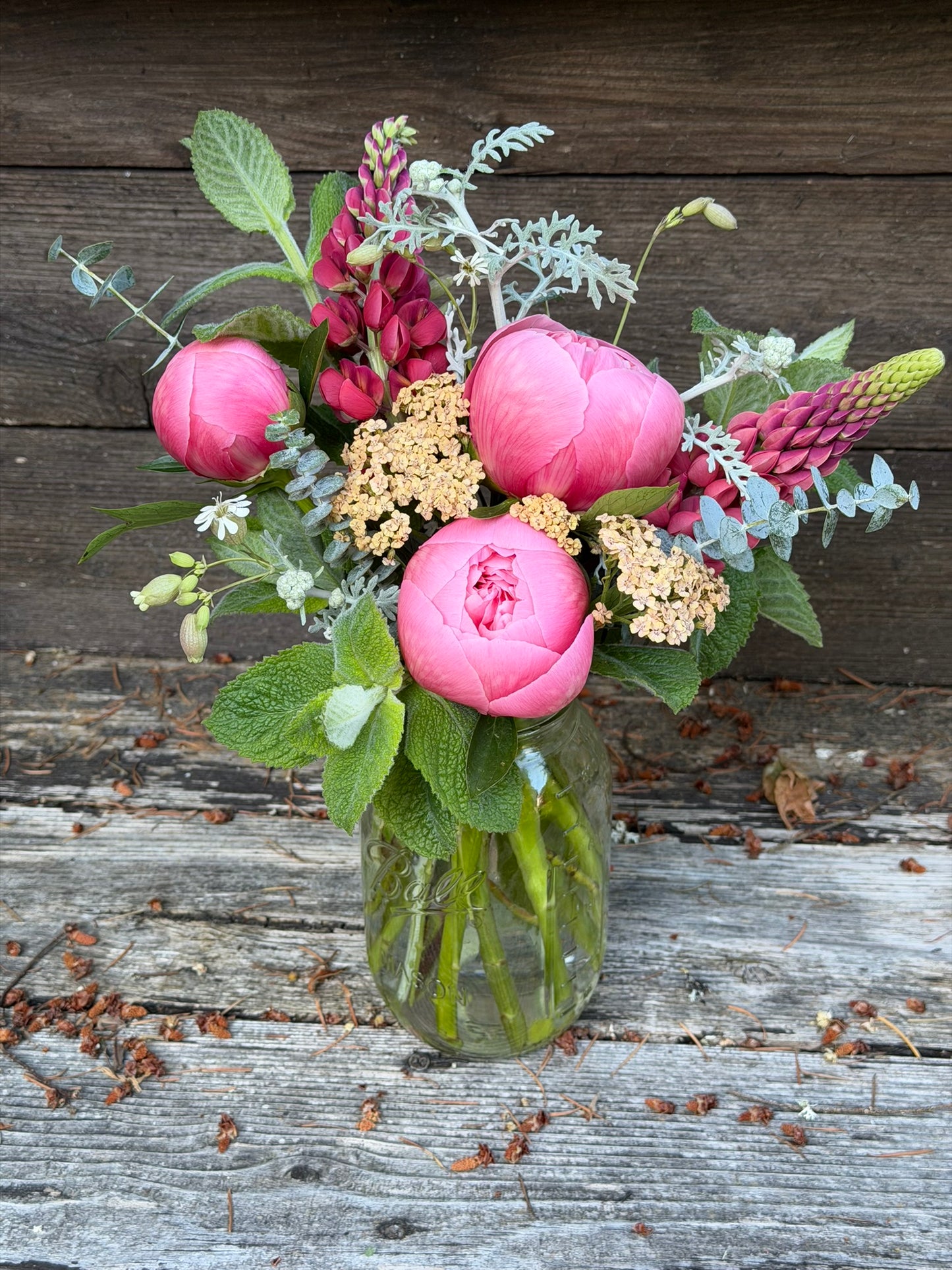 The graceful-stem Bouquets & Arrangements for Delivery features pink peonies, lupines, yellow yarrow, green foliage, and eucalyptus beautifully arranged in a mason jar on a rustic wooden backdrop.