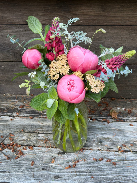 The graceful-stem Bouquets & Arrangements for Delivery features pink peonies, lupines, yellow yarrow, green foliage, and eucalyptus beautifully arranged in a mason jar on a rustic wooden backdrop.