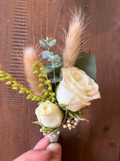 A hand holds The Graceful Stem Boutonniere with white roses, green leaves, white buds, and dried grasses against wood—a romantic floral accessory ideal for prom or online boutonniere delivery.