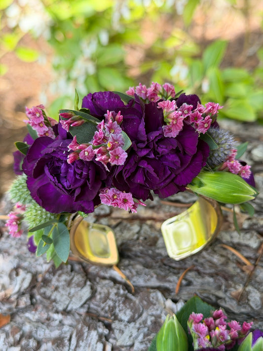The Graceful Stem's Corsage is a gold bracelet decorated with deep purple and pink flowers, beautifully displayed on bark outdoors—a charming handmade piece ideal for weddings or proms.