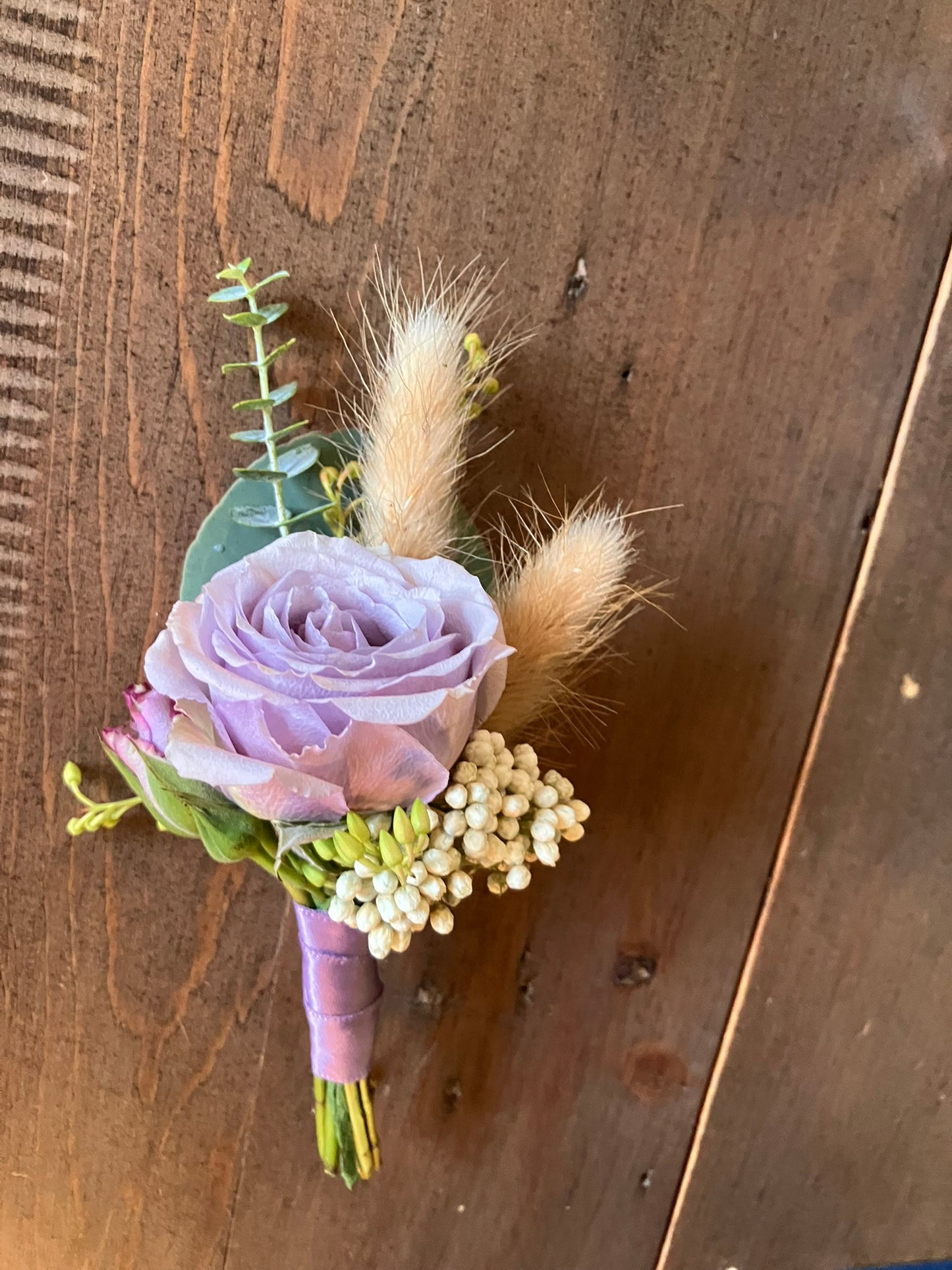 The Graceful Stem's Boutonniere features a lavender rose, greenery, and dried bunny tails, arranged in a romantic floral style and displayed on a wooden surface.