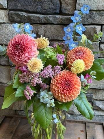 A large Graceful Garden Vase by The Graceful Stem holds an assortment of seasonal blooms—dahlias, chrysanthemums, and assorted greenery with small blue, pink, and yellow flowers—artfully displayed against a stone wall background.