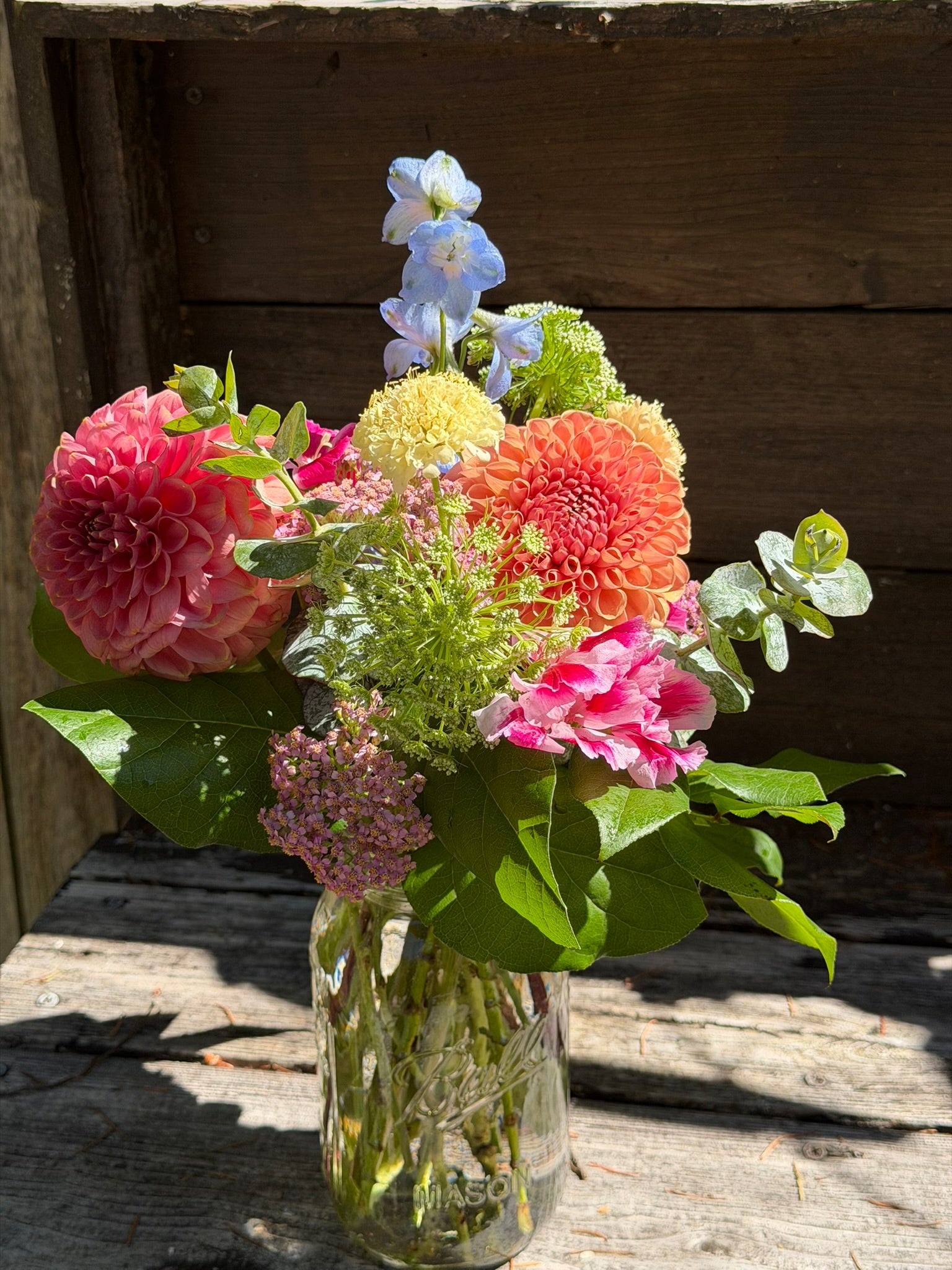 A Graceful Mason by The Graceful Stem, featuring a seasonal floral arrangement of assorted colorful flowers and greenery, sits on a rustic wooden surface against a wooden background.