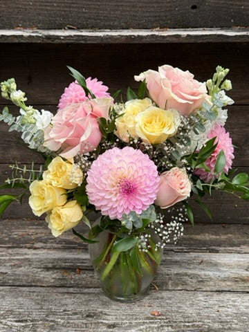 The “Gracefully Pink - Classic” by The Graceful Stem features a charming glass vase filled with pink dahlias, pale yellow and pink roses, white baby’s breath, and assorted greenery. Set on a rustic wooden surface with a wooden background, this lovely arrangement is perfect for enhancing your October floral campaign or as thoughtful breast cancer awareness flowers.