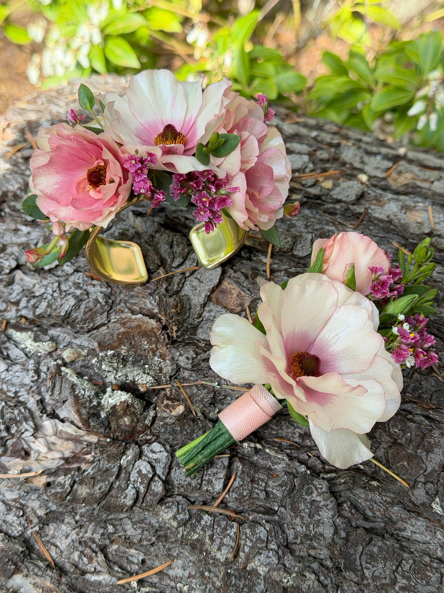 The graceful-stem Corsage, featuring pink and white blooms on a gold bracelet with a matching boutonniere, rests on tree bark amid lush greenery.
