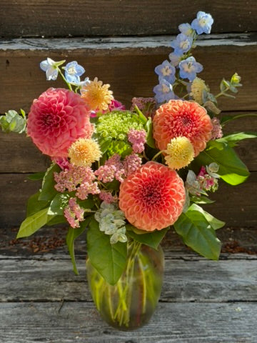 The Graceful Garden Vase by The Graceful Stem showcases a lush arrangement of dahlias, delphiniums, and assorted greenery in their signature style, with seasonal blooms beautifully displayed against a rustic wooden background.