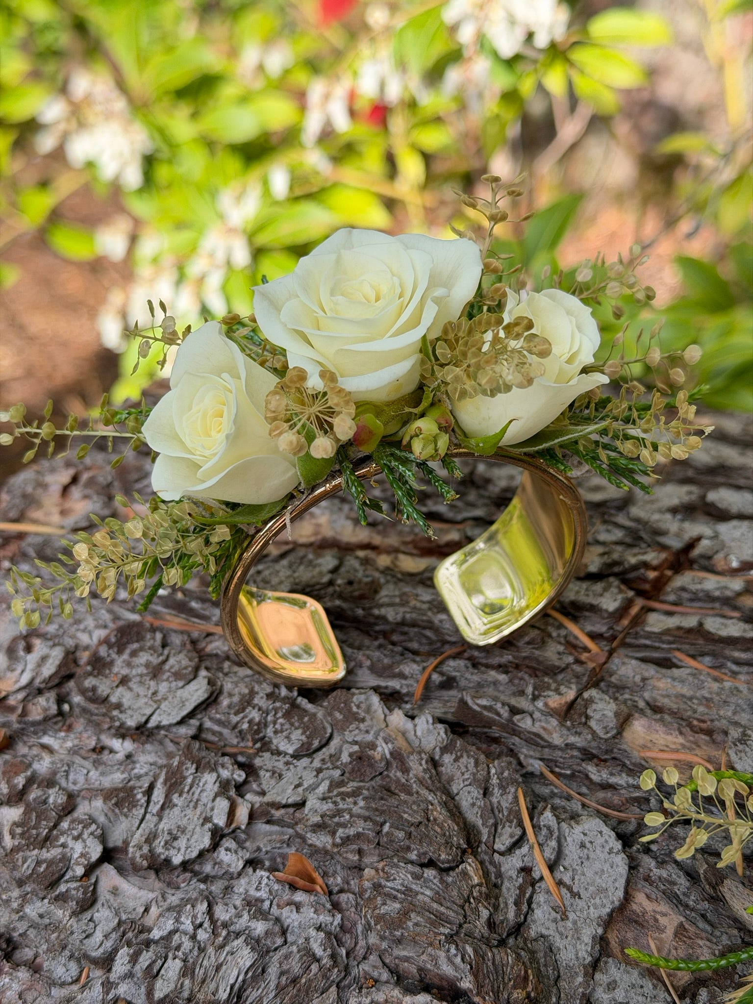 The Graceful Stem’s Corsage is a handmade gold cuff bracelet adorned with three white roses and greenery, showcased on tree bark with vibrant foliage in the background.