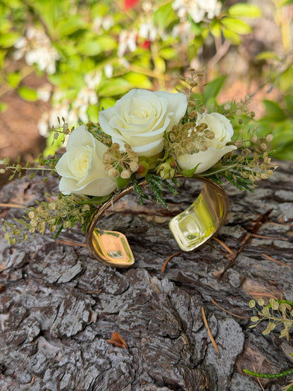 The Graceful Stem’s Corsage is a handmade gold cuff bracelet adorned with three white roses and greenery, showcased on tree bark with vibrant foliage in the background.