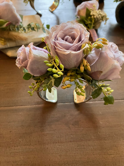 The Graceful Stem’s Corsage features three pale purple roses with greenery in a silver holder, displayed on a wooden table.