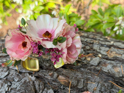 The Graceful Stem's Corsage gold cuff bracelet, adorned with light pink and purple flowers, is displayed on textured tree bark with green foliage in the background.