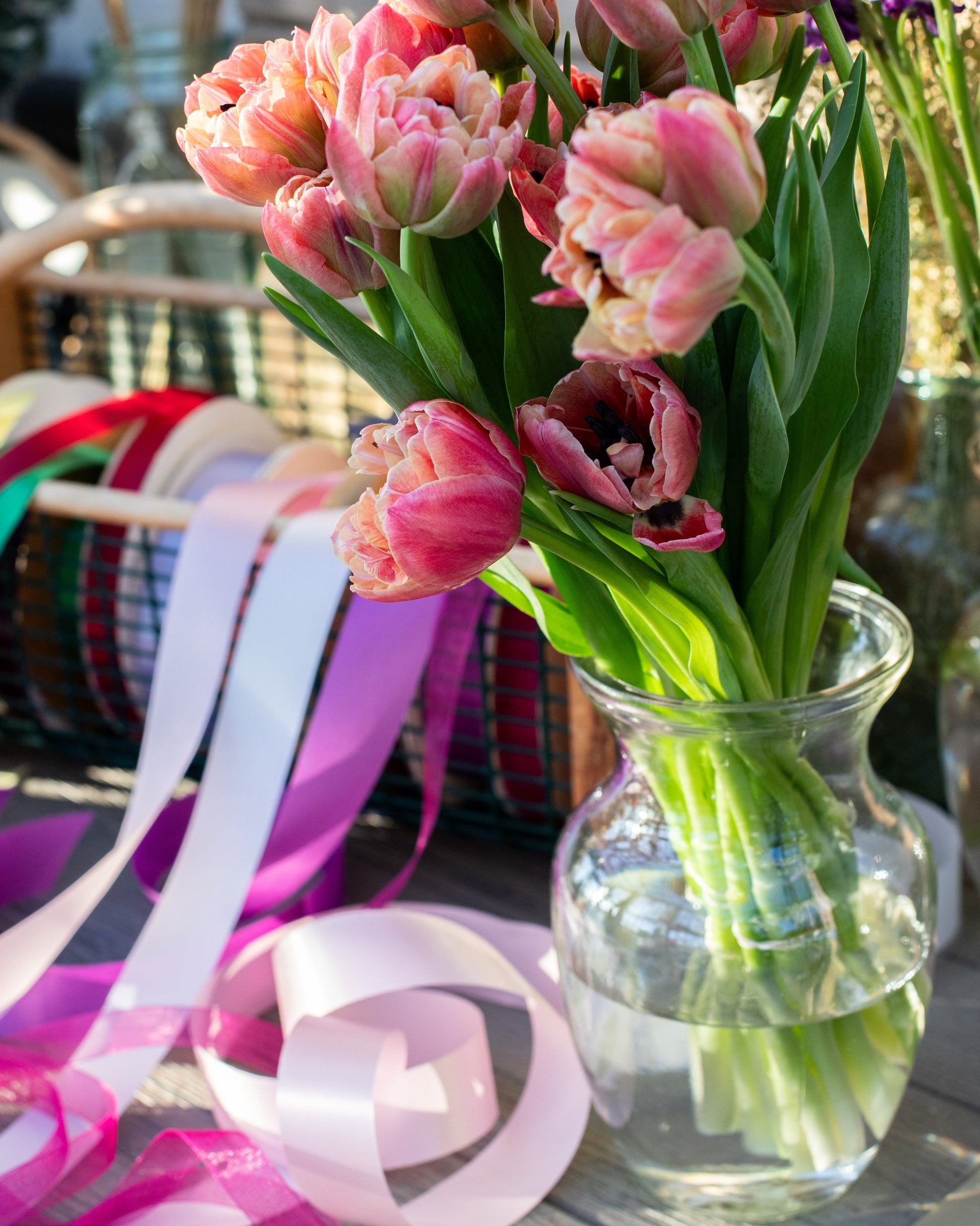 The Graceful Stem’s Flower Bar features a glass vase with pink tulips, colorful ribbons, and a basket on a sunlit table—creating a charming floral display.