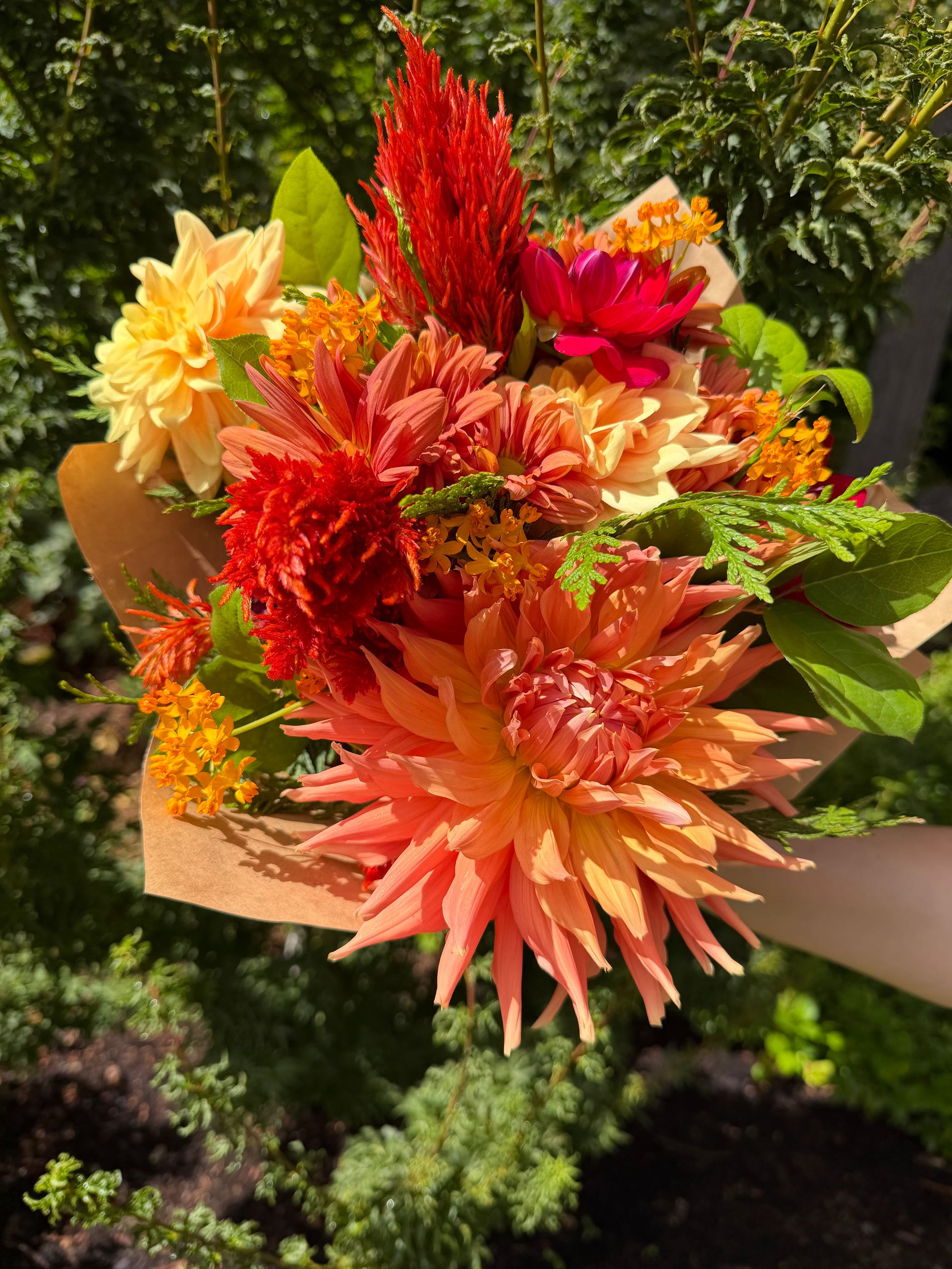 A hand holds the Gracefully Hand-Tied Bouquet by The Graceful Stem, featuring fresh dahlias and an assortment of orange, red, and yellow flowers with green leaves, set against a garden background.