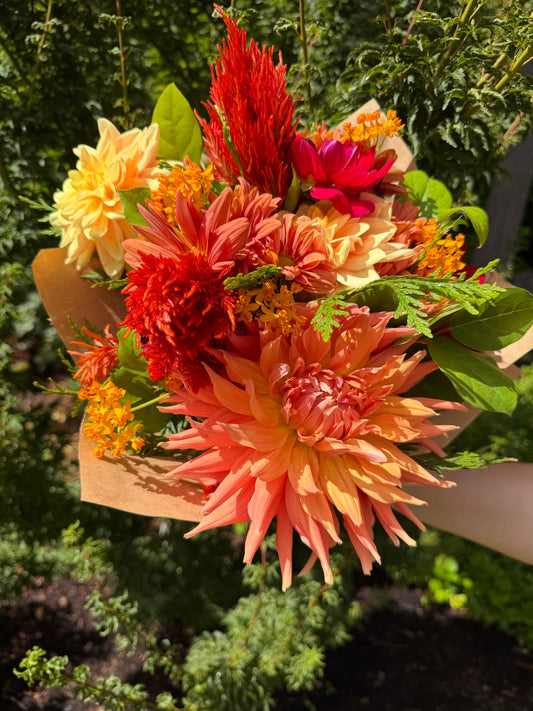 A hand holds the Gracefully Hand-Tied Bouquet by The Graceful Stem, featuring fresh dahlias and an assortment of orange, red, and yellow flowers with green leaves, set against a garden background.