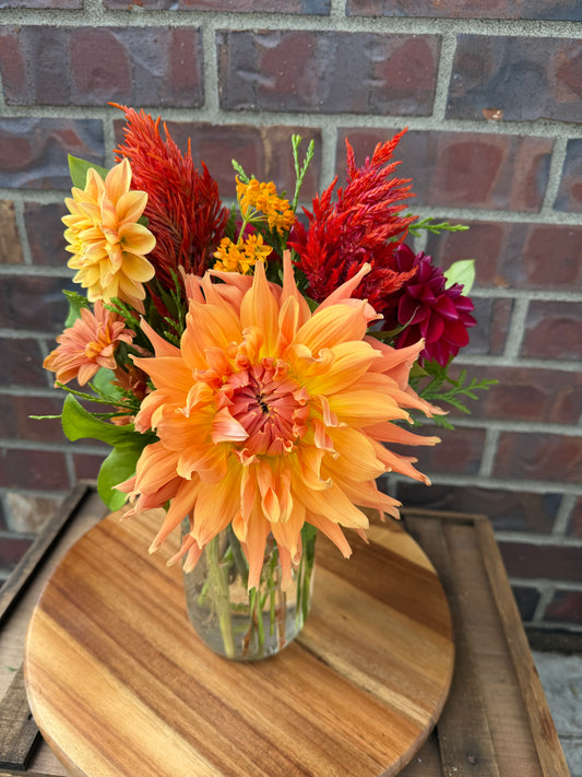 A Graceful Mason glass jar by The Graceful Stem holds a beautiful floral arrangement of orange, yellow, and red flowers, displayed on a round wooden board in front of a brick wall.