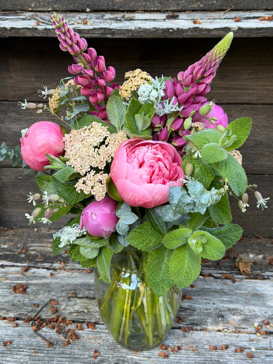 The Graceful Garden Vase Bouquet by The Graceful Stem features pink peonies, purple lupines, yellow yarrow, and assorted greenery in a glass vase, beautifully arranged on a rustic wooden surface.