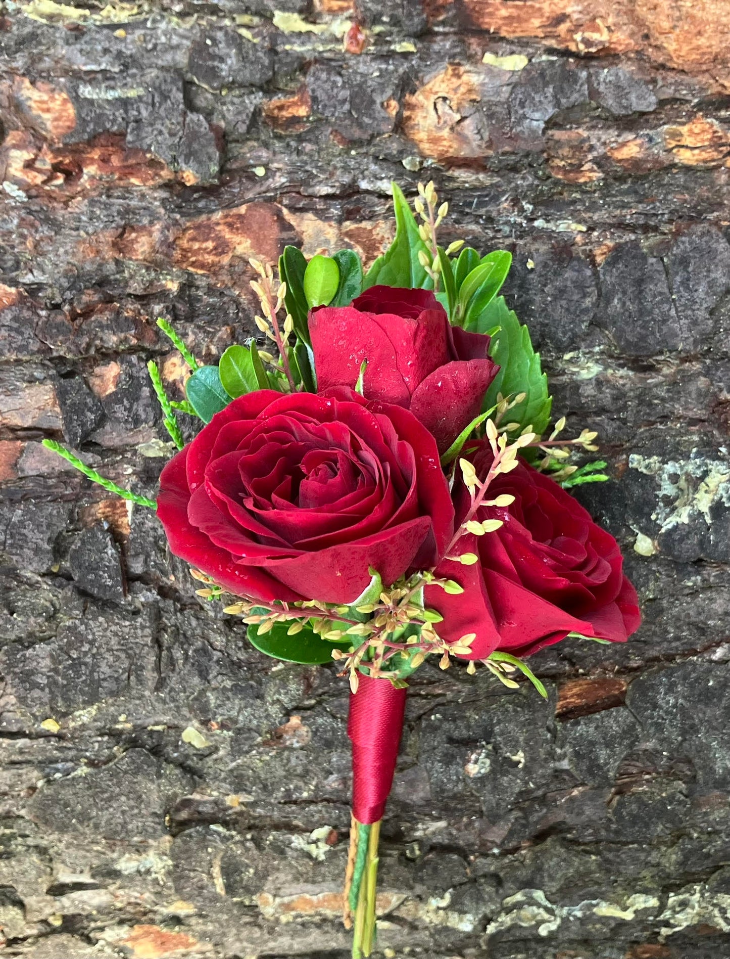 The Graceful Stem's Boutonniere features a romantic design with three red roses, greenery, and yellow buds wrapped in red ribbon—perfect for special occasions or online delivery. Shown against a textured stone background.