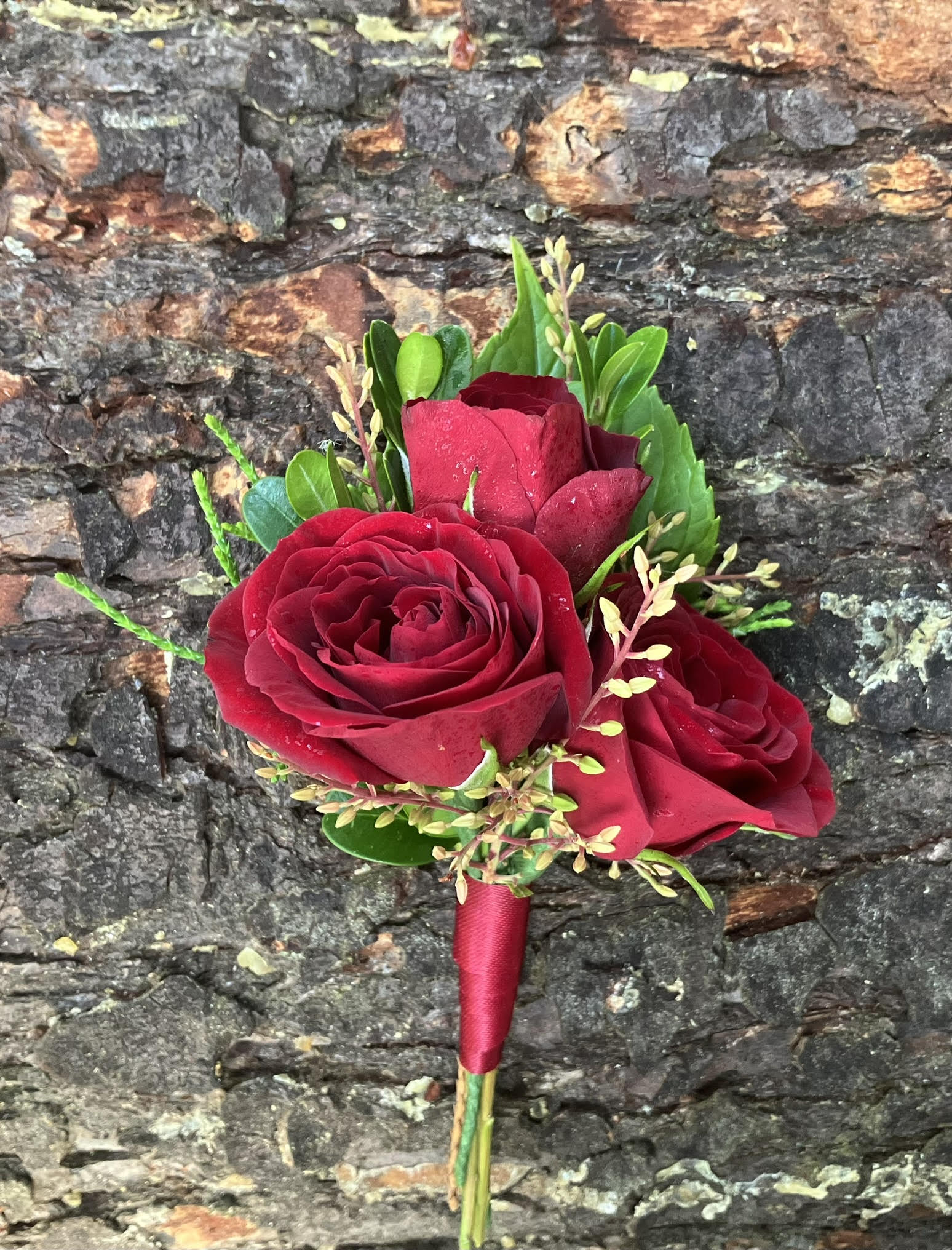 The Graceful Stem's Boutonniere features a romantic design with three red roses, greenery, and yellow buds wrapped in red ribbon—perfect for special occasions or online delivery. Shown against a textured stone background.