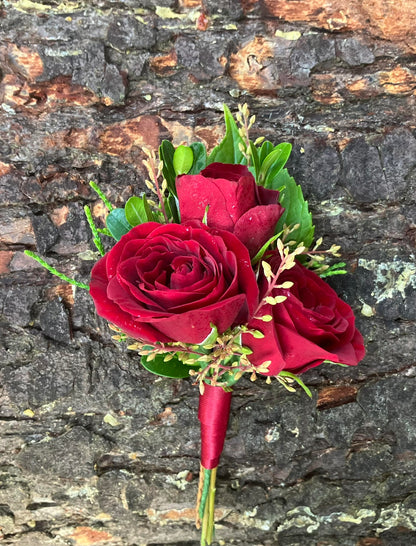 The Graceful Stem's Boutonniere features a romantic design with three red roses, greenery, and yellow buds wrapped in red ribbon—perfect for special occasions or online delivery. Shown against a textured stone background.