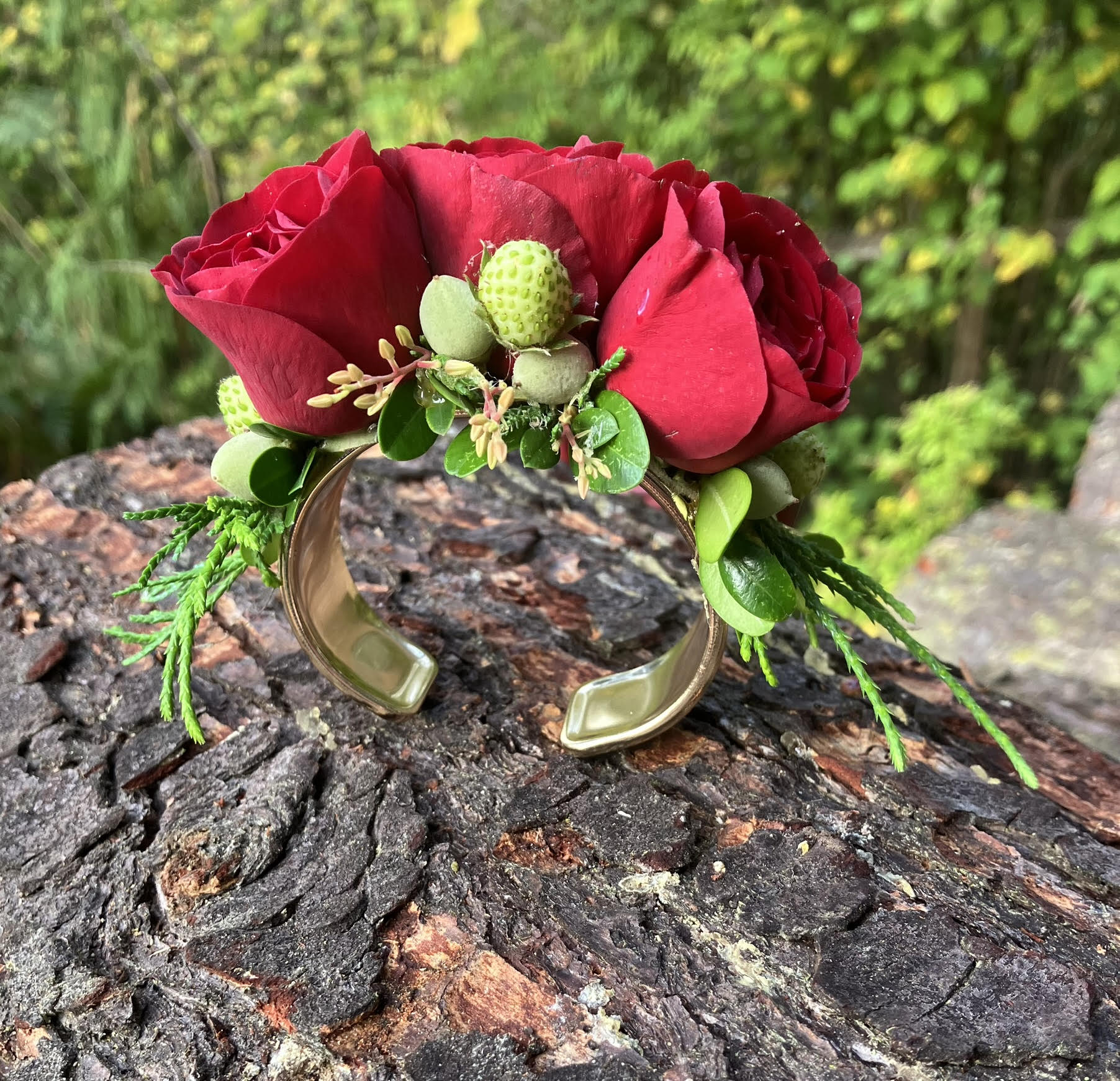 The Graceful Stem's Corsage features a gold cuff bracelet adorned with red roses, green berries, and assorted greenery—a handmade piece perfect for any occasion. Shown here displayed on tree bark with lush foliage in the background.