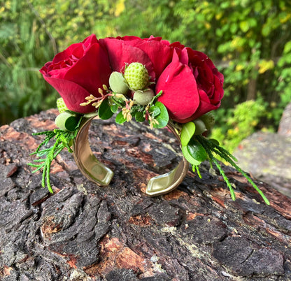 The Graceful Stem's Corsage features a gold cuff bracelet adorned with red roses, green berries, and assorted greenery—a handmade piece perfect for any occasion. Shown here displayed on tree bark with lush foliage in the background.